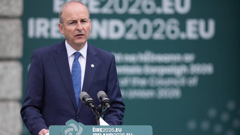 Taosieach Micheál Martin. He is wearing a navy suit and standing at a green lectern. Behind him is a large green banner with 'IRELAND2026.EU' written on it.