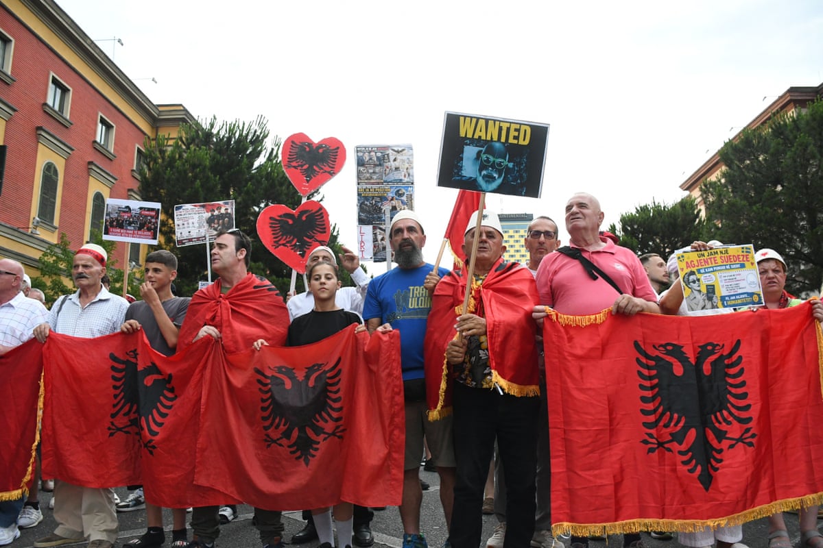 Protesters stand with red flags bearing a black double-headed eagle. Placards include one with the word: Wanted and a picture of the Albanian prime minister.
