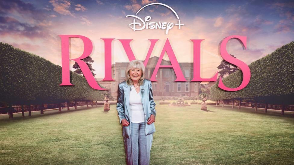 Dame Jilly Cooper wearing a blue suit and trousers. She stands in front of a