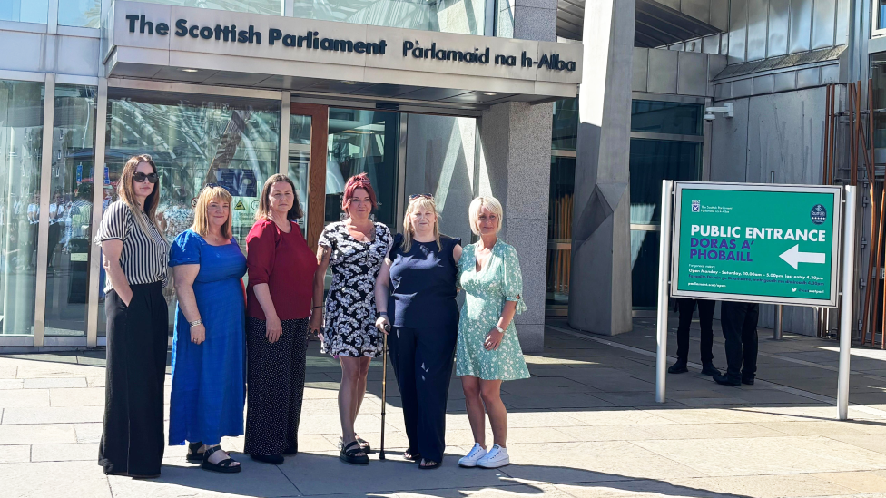 Group of six people standing outside the entrance to the Scottish Parliament building on a bright sunny day. The glass-fronted entrance displays signs reading “The Scottish Parliament” and “Pàrlamaid na h-Alba”, while a green sign for the public entrance stands to the right.