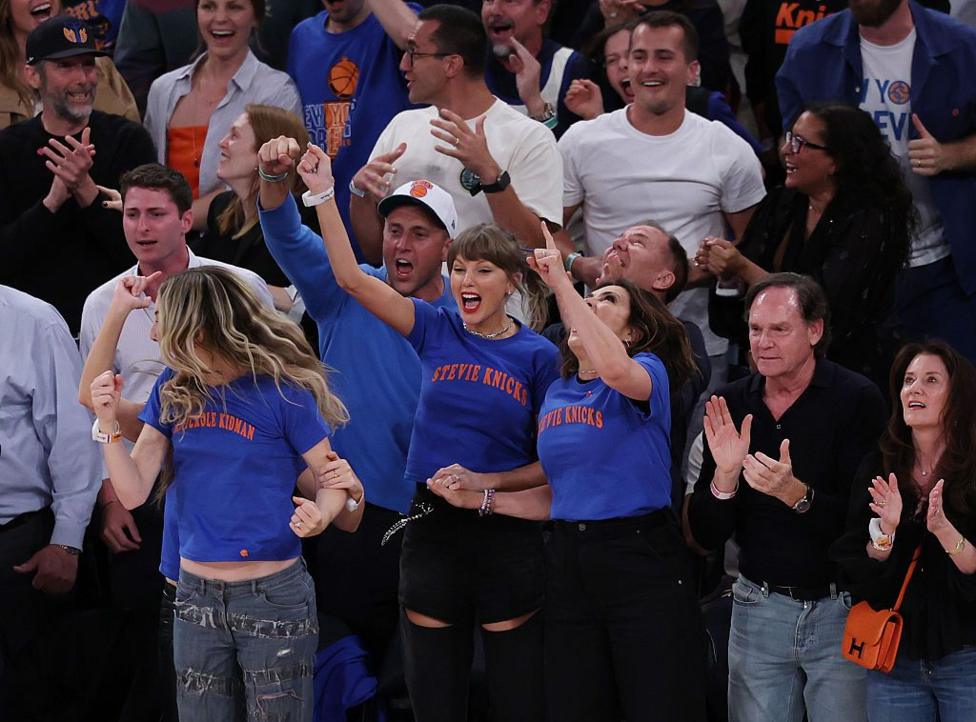 Taylor Swift and others are seen courtside cheering for the New York Knicks. She is wearing a blue T-shirt with orange lettering - the Knicks colors. 
