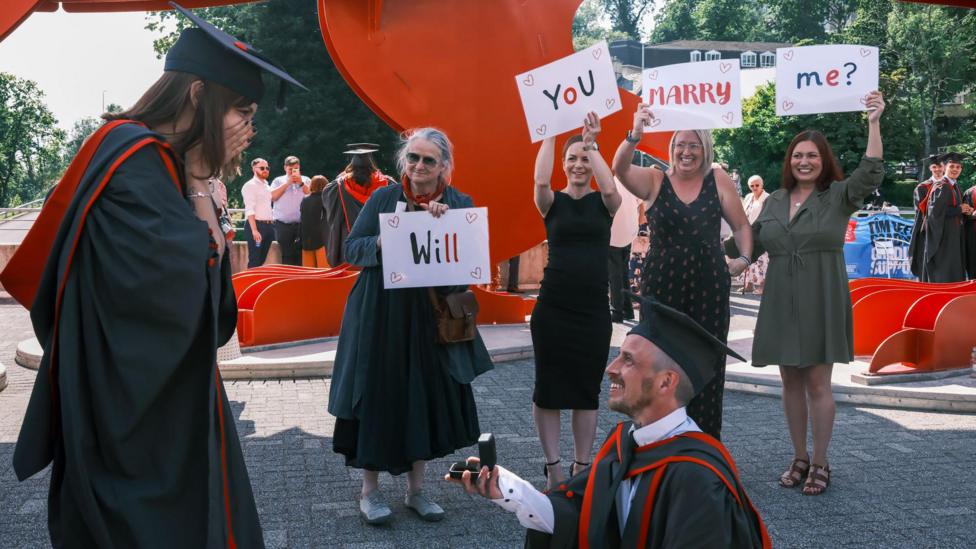 Kat standing with her hands over her mouth looking at Steve on one knee holding a ring. Four people stand behind holding signs saying 'Will you marry me?'