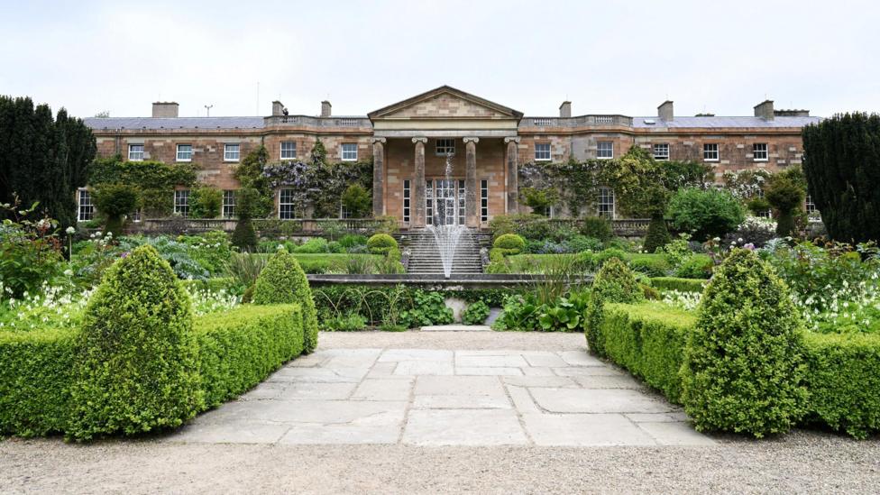 A wide view of Hillsborough Castle and its garden courtyard on a cloudy day. The castle is a large, detached two and three-storey multi-bay neoclassical house with a red sandstone ashlar exterior. A circular fountain and a set of stone steps lead up from the garden to the castle's central portico. The landscaped courtyard contains neat box hedging and tall Irish yew trees among other plants and shrubs.