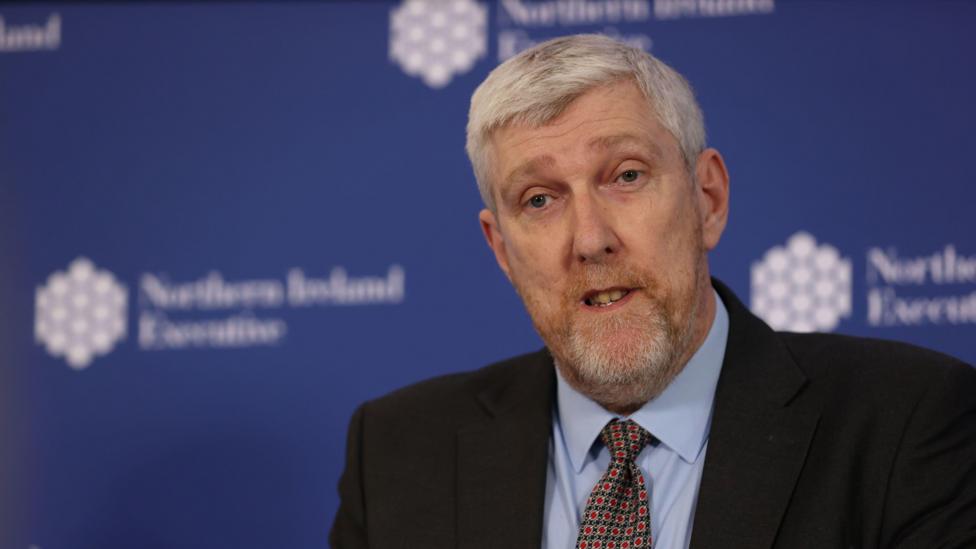 Finance Minister John O'Dowd, a man with short grey hair, speaking to the media at a previous press conference. He is wearing a black suit, blue shirt and red, yellow and navy tie. He is standing in front of a plain navy background which is decorated with a