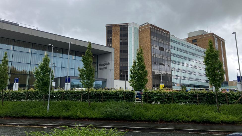 A wide shot of Altnagelvin Hospital on a cloudy day. It is a tall, multi-story building with brown brick, glass and other materials featured in its front elevation. The main entrance features a tall, glass-fronted atrium attached to the main tower block. Hedges and trees line the road outside the building.