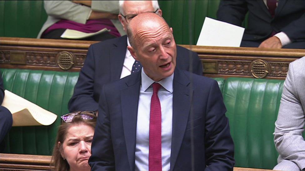 A middle aged man with a bald head, wearing a navy blue suit over a light blue shirt and burgundy tie, stands on the green benches of the House of Commons holding notes in his hand. 