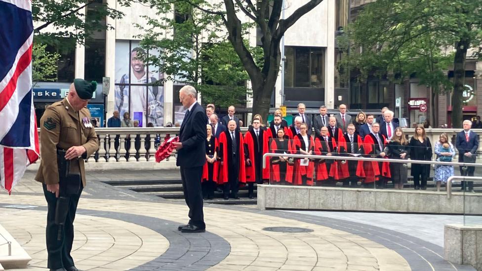 To the left there is a union flag. An armed forces representative dressed in a brown military uniform bowing his head. In the centre of the photo is a man with a dark suit and grey hair, who is holding a poppy wreath. In the background there are people dressed in red gowns lined up watching the ceremony.