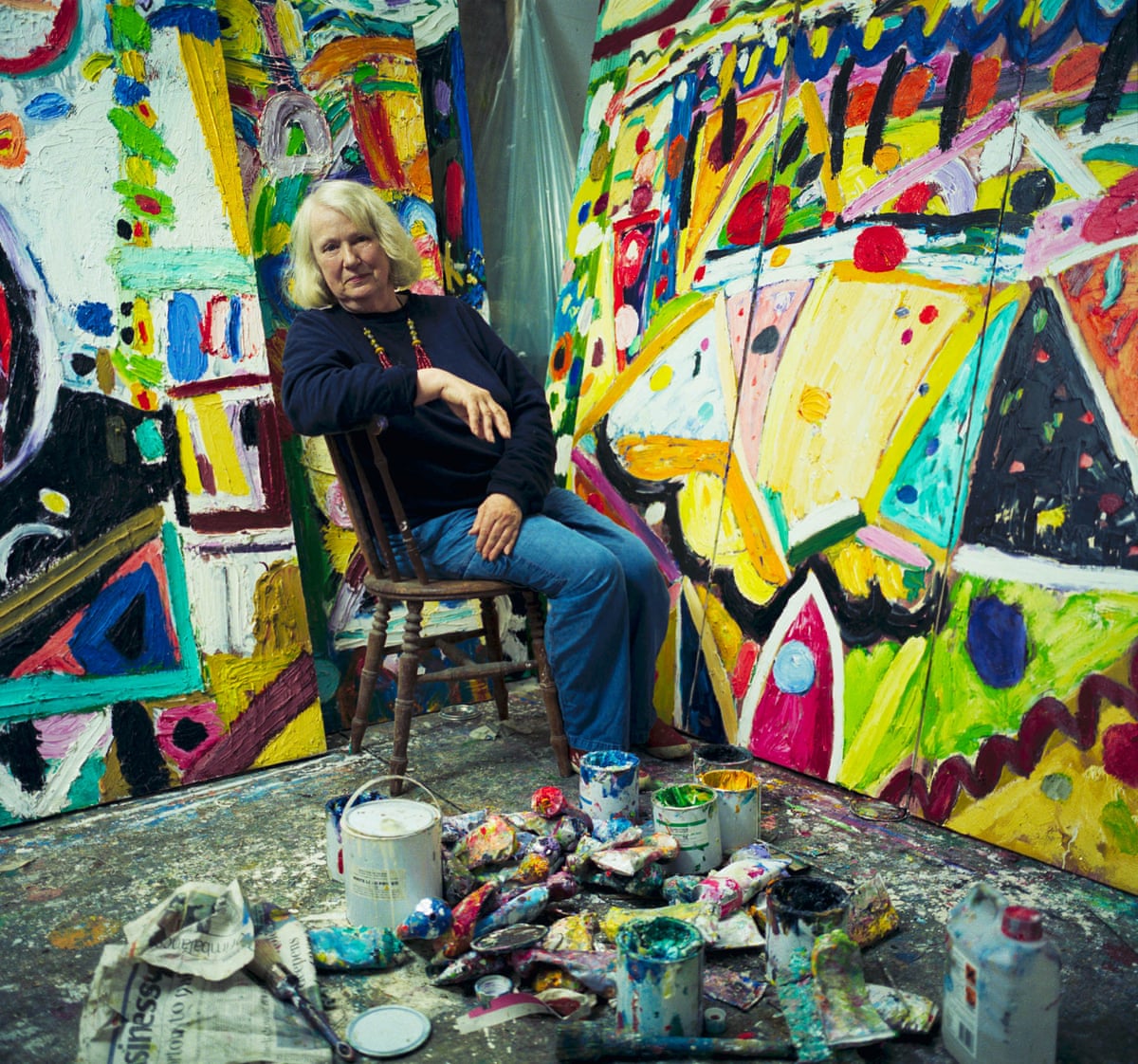Wide shot of Ayres sat beside bold, large canvas, with paints and cans strewn across studio floor
