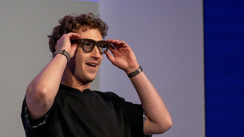 Mark Zuckerberg, wearing a black t-shirt and smiling, interacts with a pair of Meta Ray-Ban Display glasses in front of an audience.