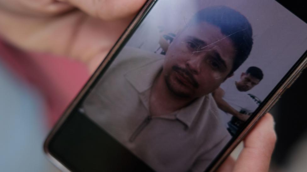 A phone displays the photo of a man in a beige zip up sweater