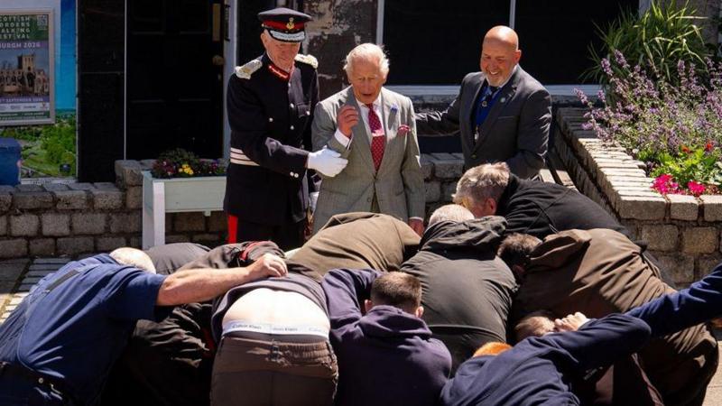 King Charles III is held back by a man in uniform and a man with a bald head as a group of men grapple for a ball in front of him