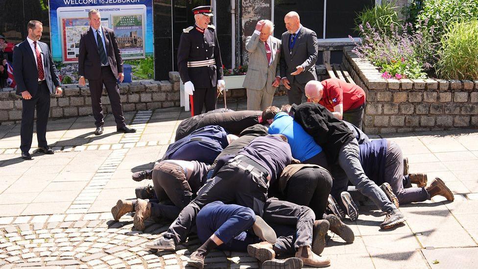 A scrummage of men on the ground in front of five men standing in suits