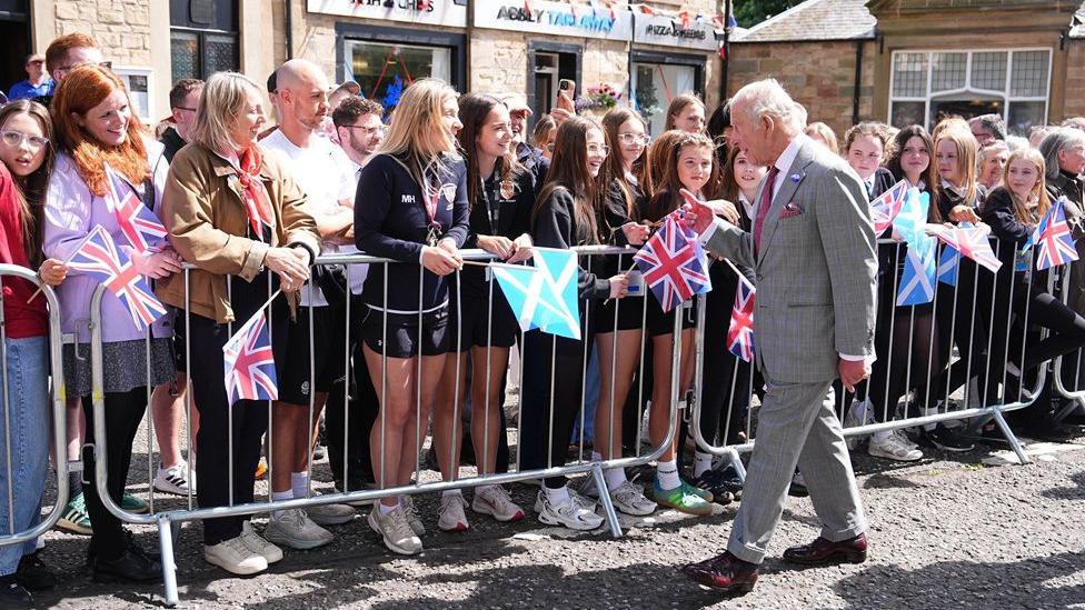 A big gathering of people waving flags to greet King Charles