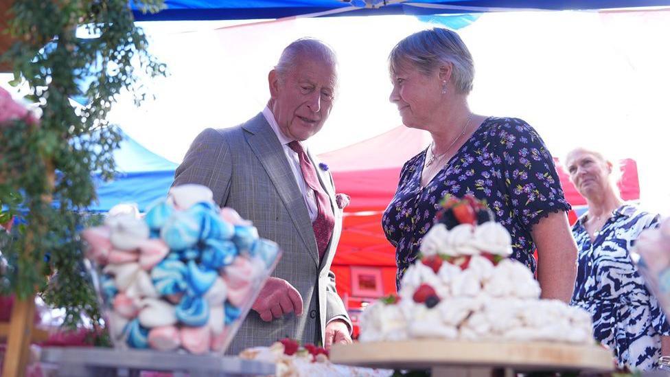 King Charles talks to a woman in a patterned dress in front of a tower of meringues