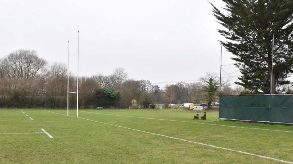 An empty rubgy field with a goal and white lines painted on the grass. There is a green fence to the right, and trees and parked cars in the background. The sky is grey.