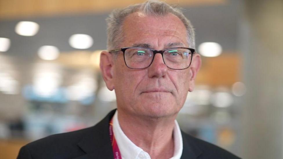 A head and shoulders shot of a man with short white hair, wearing black-framed glasses and a dark jacket over a white shirt. He is inside.