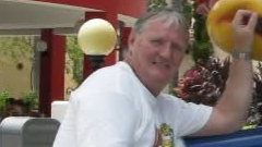 A slightly blurry picture of a man in his 60s with grey short hair, wearing a white t-shirt. He has his one arm raised in the air. 