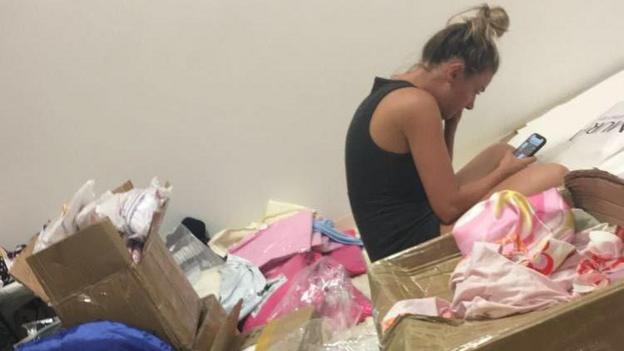 Olivia hunched over her phone with her hair in a bun surrounded by boxes of clothes.