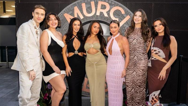 A man on the left stands next to six women in front of a sign saying 'MURCI' . He wears a cream linen suit, while the women all wear maxi dresses of different colours and patterns.