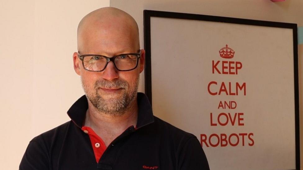 Ingmar Posner in glasses and a black polo shirt stands in front of a pictures that reads: