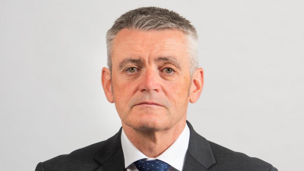 A man with short grey hair looks into the camera wearing a black suit with white shirt and navy tie. He sits in front of a white background.