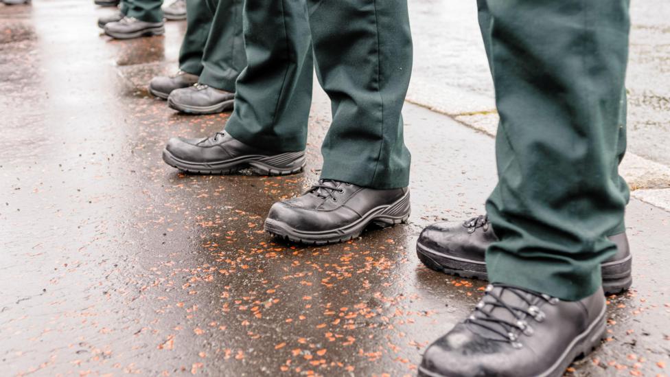 Police officers' boots as they stand in line. They are outside and the pavement is wet. The officers are wearing black boots with laces and dark green trousers. 