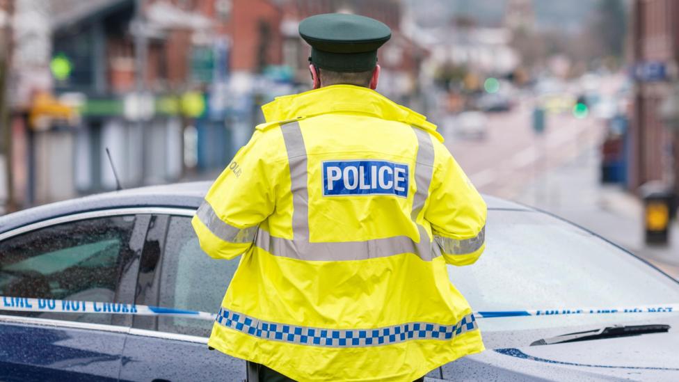 A PSNI police officer wearing a high-visibility jacket closes off a street. Blue and white police tape is up. The street in the background is out of focus.
