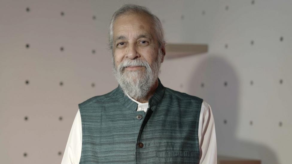 Madan Lokur is stood in a room with white walls, looking at the camera. He is wearing a sleeveless jacket over a white top and has grey hair and a grey beard.