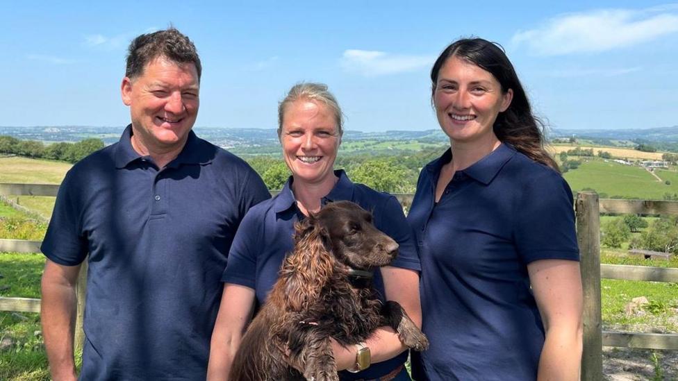 A man with short brown hair is wearing a plain blue t-shirt. He is smiling and standing next to two women wearing the same coloured shirts. The woman in the middle is holding a brown dog. 