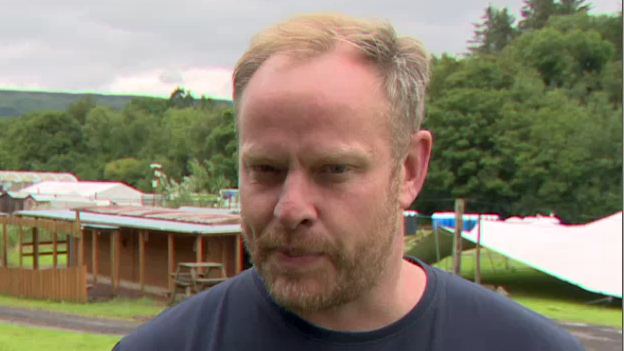 Ross Parkhill has short blonde hair and is wearing a navy shirt and is standing near a festival camping site.