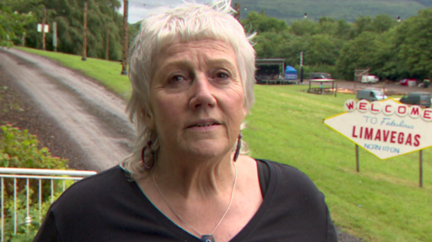 Diane Greer has long white hair and is wearing a black top and silver necklace. She is standing in the festival grounds.
