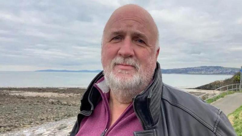 David Graves. He has a bald head and short grey beard. He is wearing a black jacket, perhaps made from leather, with a purple zip up top underneath