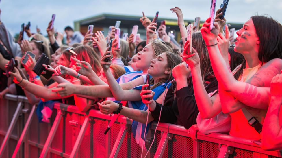 A crowd of young people at the barrier at an open-air gig. Many of them are filming a performance on their phones. There is a red tinge to the photo