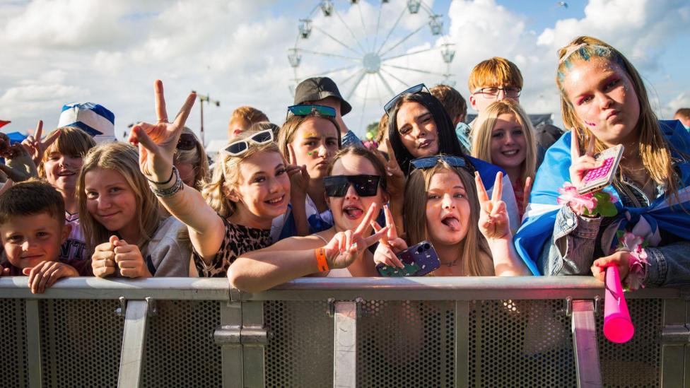A crowd of young people behind a barrier at a sunny outdoor music festival.