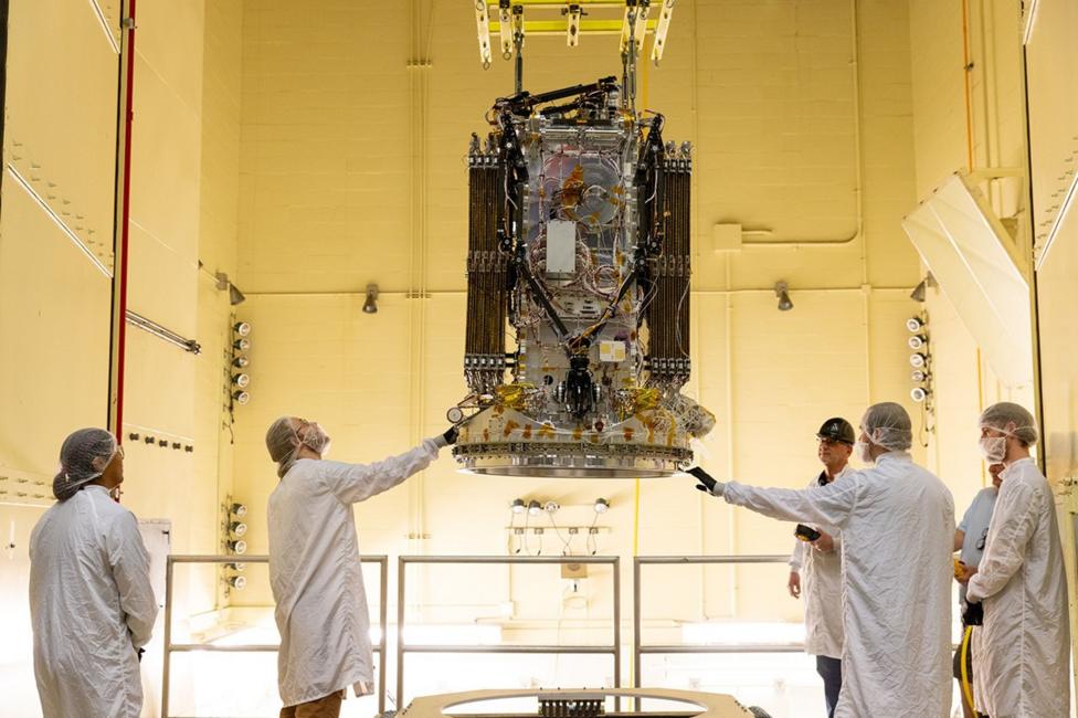 In a bright yellow test chamber, six engineers in white lab coats and hairnets stand around a large, boxy spacecraft suspended from the ceiling. They reach up with tools and gloved hands, examining its exposed wires, pipes, and shiny metal panels during a precision vibration test.