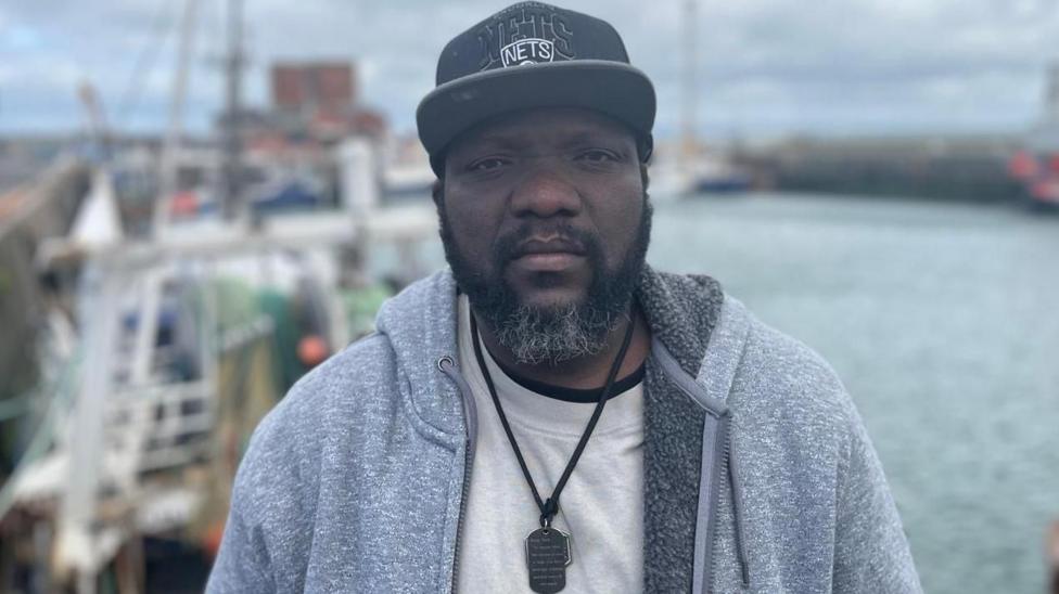 Gershon Norvivor is wearing a grey fleece hoodie, white T-shirt and black baseball cap. He is staring at the camera with a serious expression on his face. He has a black beard and a moustache.