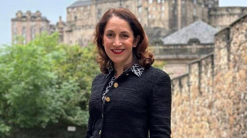 Katerina Brown has shoulder-length, brown hair. She is smiling while standing in front of Edinburgh Castle. She is wearing a black and white shirt and a black jacket