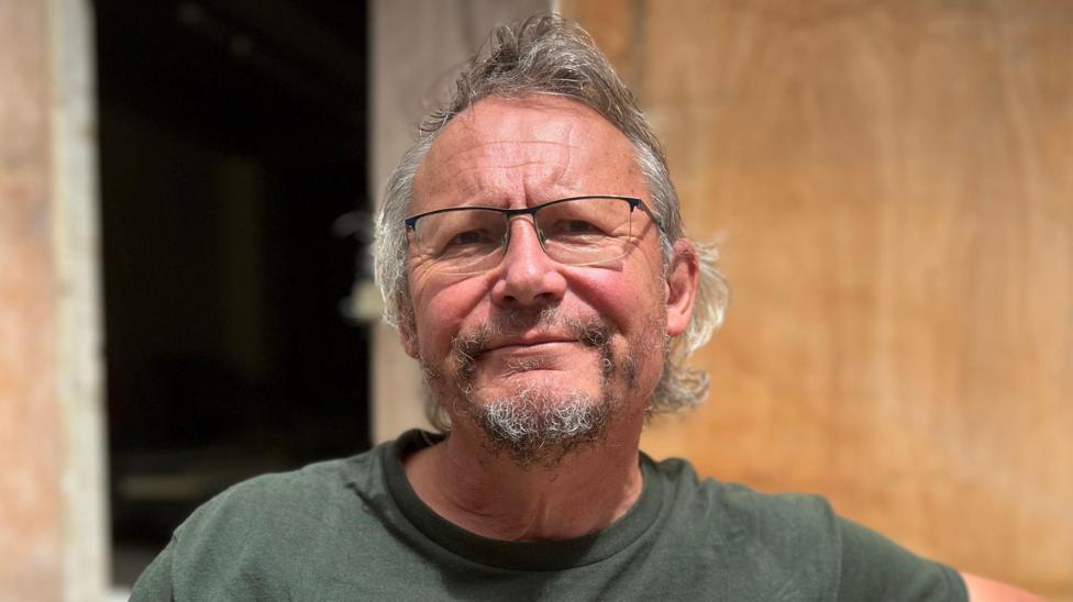 Mark is wearing glasses and a dark green graphic T-shirt sits on a wooden bench outdoors. The background features a beige stone wall and a doorway, with bright daylight illuminating the scene.