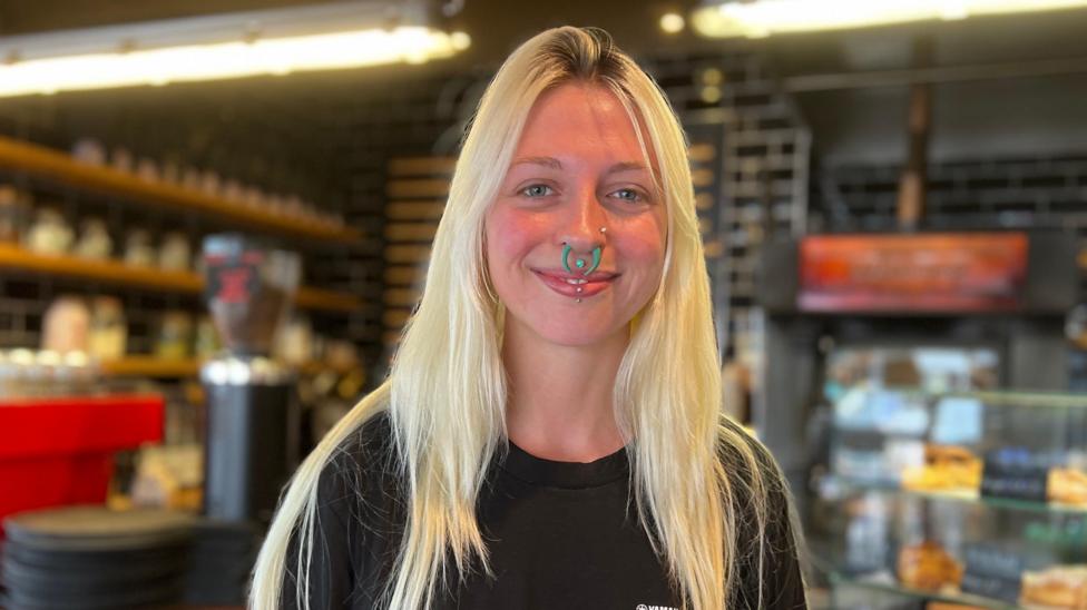 Sarah has long light-coloured hair stands behind the counter of a café, wearing a black T-shirt and facial piercings. Shelves lined with cups and supplies are visible in the softly blurred background, alongside a glass display case of baked goods