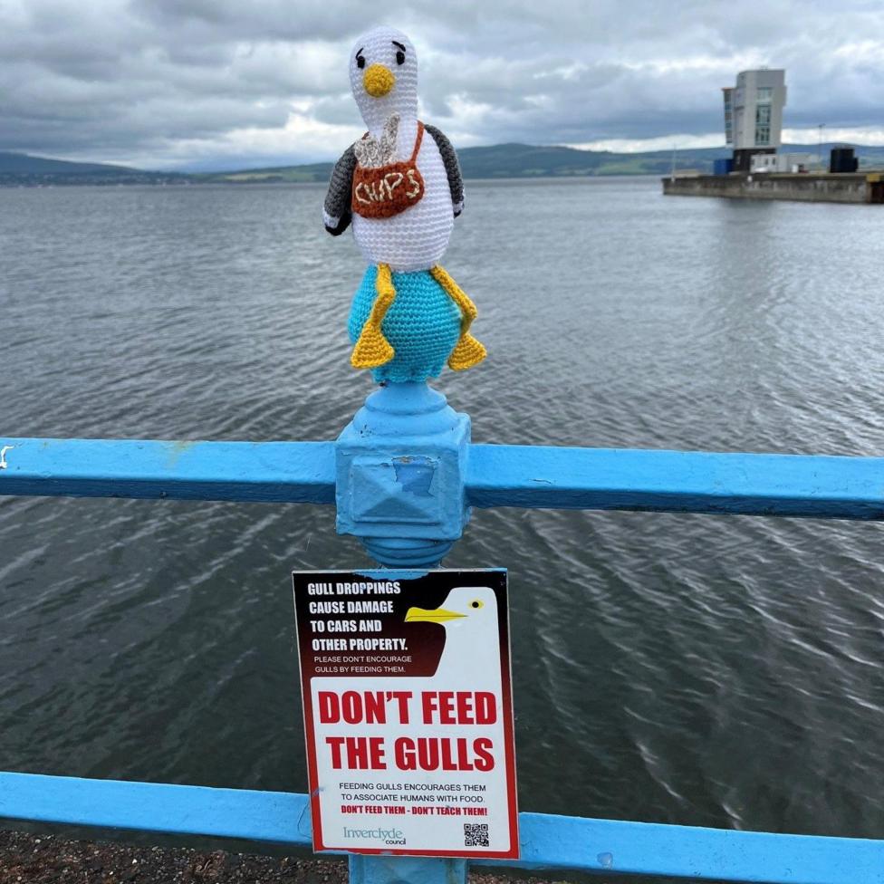A knitted seagull figure holding a bag labelled “CHIPS” is perched on a blue waterfront railing above a sign reading “DON’T FEED THE GULLS.” The scene overlooks a wide estuary with hills and a cloudy sky in the background.