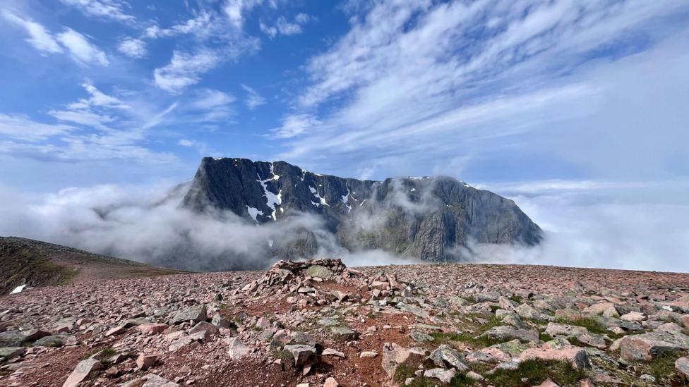 A dramatic mountain landscape showing rugged, rocky peaks rising through low cloud and mist, with patches of lingering snow beneath a bright blue sky streaked with thin clouds.