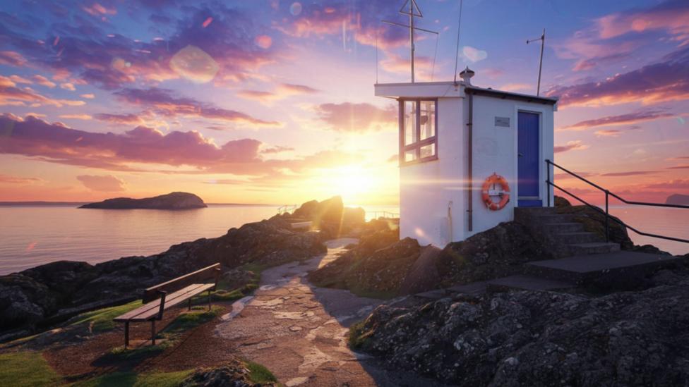 A small, white harbour lookout building stands on a rocky shoreline at sunset. Bright sunlight and colourful clouds illuminate the scene, with a bench, coastal path, and offshore island visible in the background.