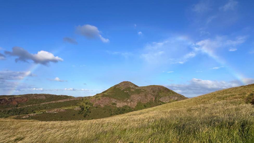 A grassy hillside rises toward a rugged green-and-brown hill beneath a bright blue sky. Faint rainbow arcs are visible on either side of the landscape, with scattered clouds adding depth to the scene.