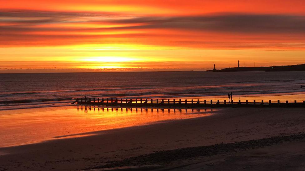 A vivid red and orange sunrise spreads across the horizon above a sandy beach. Reflections of the colourful sky shimmer on the wet sand, while a groyne and distant lighthouse stand in silhouette against the sea.