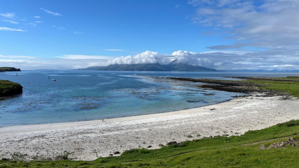 A sweeping coastal landscape featuring a white sandy beach, crystal-clear blue water, and distant mountains partly draped in clouds under a bright sky.