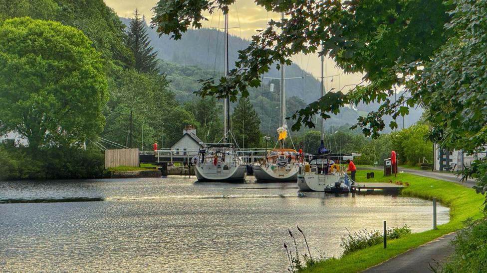 A peaceful canal scene featuring several moored sailboats near a lock, framed by lush green trees and rolling hills under a cloudy, golden-lit sky.
