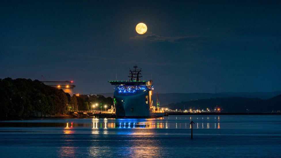 A large vessel moored on calm water at night beneath a bright full moon, with colourful dock lights reflecting across the water’s surface.