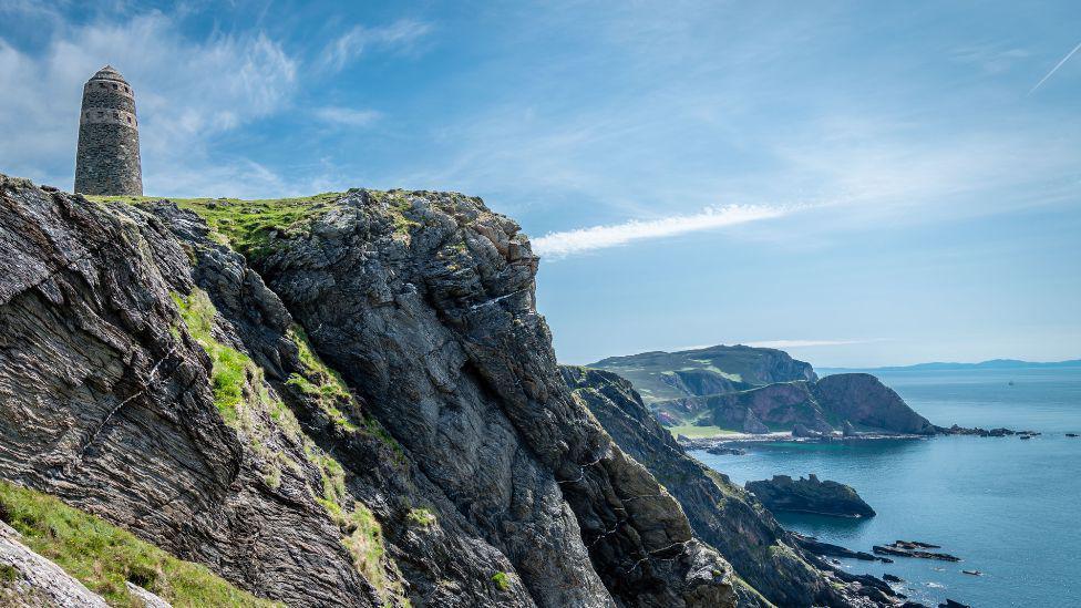 Dramatic sea cliffs rising above a rugged shoreline, topped by a solitary stone tower overlooking the ocean and distant headlands beneath wispy clouds.