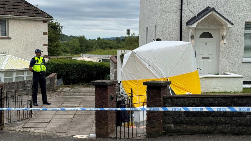 The scene at Rocky Road, Merthyr Tydfil. There's a forensic tent outside a white house with a brown brick wall. An officer is stood on the left hand side and there's police tape in front of the house.