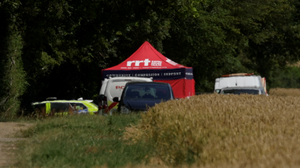Vehicles belonging to the police and other emergency workers parked by the side of the field, where a red tent has been set up.